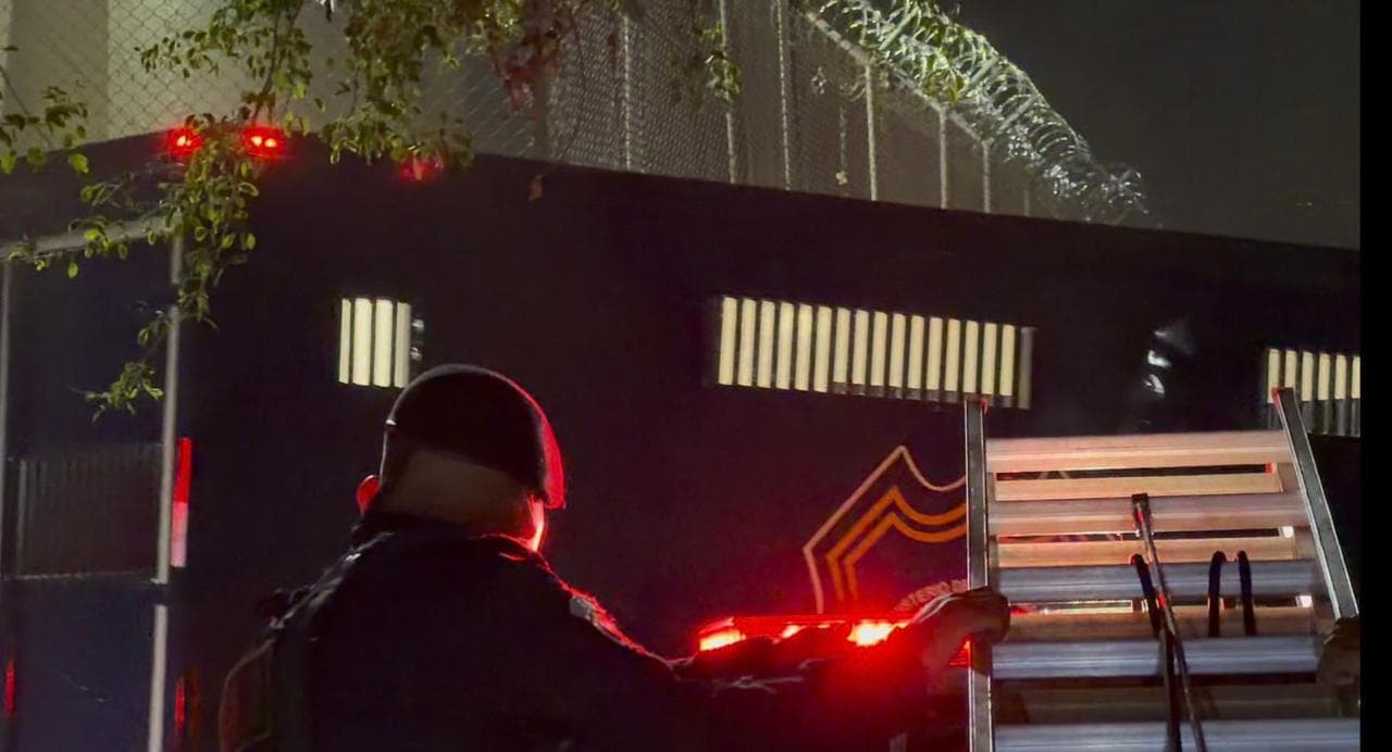 Exterior nocturno de un centro penitenciario, con reja perimetral y agente de seguridad